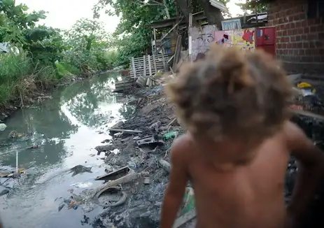 Arquivo/Fernando Frazão/Agência Brasil Imagem de riacho com falta de saneamento básico e uma criança passando em favela do Complexo da Maré